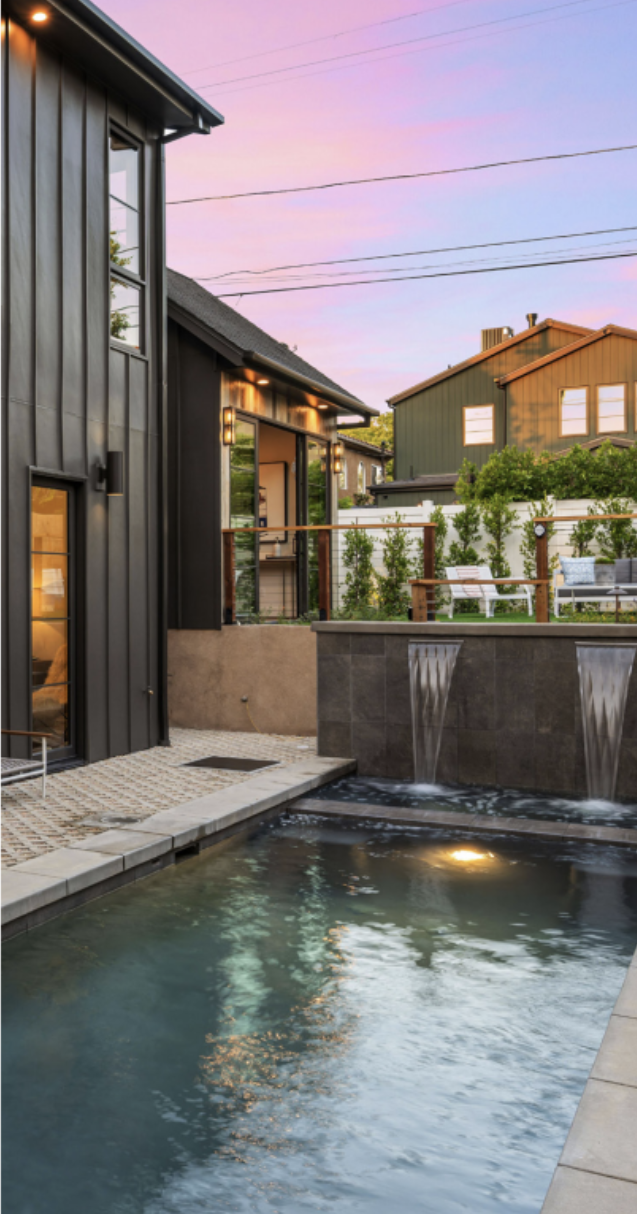 Contemporary backyard with a narrow plunge pool, cascading water features, and modern black exterior walls at sunset.